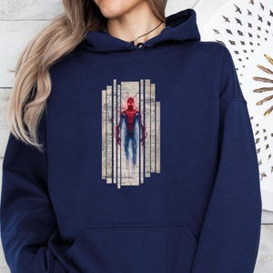 K&ouml;nnte beinhalten: Marineblauer Kapuzenpullover mit einer Grafik eines Superhelden in einem rot-blauen Anzug. Der Superheld ist in einer stilisierten, k&uuml;nstlerischen Weise dargestellt, vor dem Hintergrund vertikaler, gealterter papierartiger Tafeln. Der Hoodie hat eine Vordertasche und eine Kapuze mit Kordelzug.