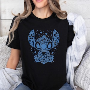 Puede incluir: Camiseta negra con un dise&ntilde;o floral azul de Stitch. El dise&ntilde;o incluye patrones florales, corazones y un personaje Stitch estilizado. La camiseta tiene cuello redondo y parece estar hecha de un material suave.
