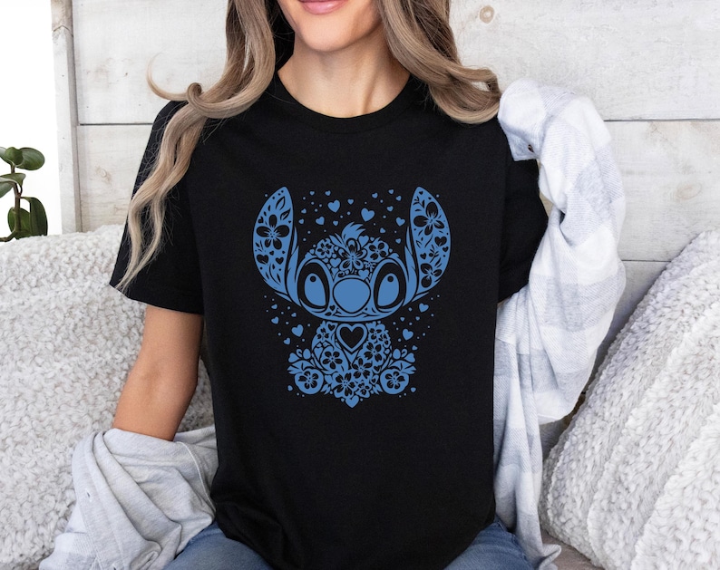 Puede incluir: Camiseta negra con un dise&ntilde;o floral azul de Stitch. El dise&ntilde;o incluye patrones florales, corazones y un personaje Stitch estilizado. La camiseta tiene cuello redondo y parece estar hecha de un material suave.