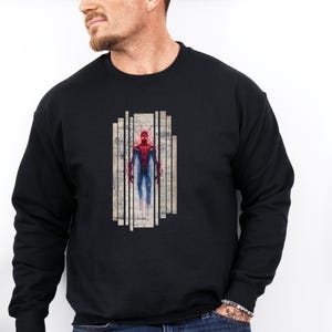 Op de afbeelding: Zwart sweatshirt met lange mouwen met een afbeelding van een superheld in een rood en blauw pak. De superheld is afgebeeld in een rechthoekig ontwerp met verticale lijnen en een vintage papieren achtergrond. De sweatshirt is gemaakt van zacht materiaal.