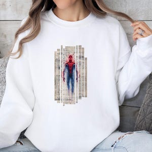 Op de afbeelding: Witte sweatshirt met een Spiderman-afbeelding. De afbeelding toont Spiderman in rood en blauw, tegen een achtergrond van vintage papieren stroken. De sweatshirt is effen wit van kleur.