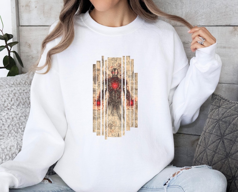 May include: White sweatshirt featuring a graphic of Iron Man in a vintage style. The design includes a detailed illustration of the character with red and brown tones, set against a background of aged paper.