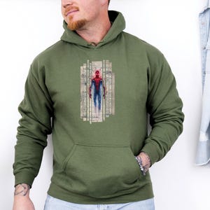 K&ouml;nnte beinhalten: Olivgr&uuml;ner Kapuzenpullover mit einer Vordertasche. Die Grafik auf der Vorderseite zeigt eine Aquarellillustration eines Superhelden in einem rot-blauen Anzug, vor dem Hintergrund architektonischer Baupl&auml;ne.