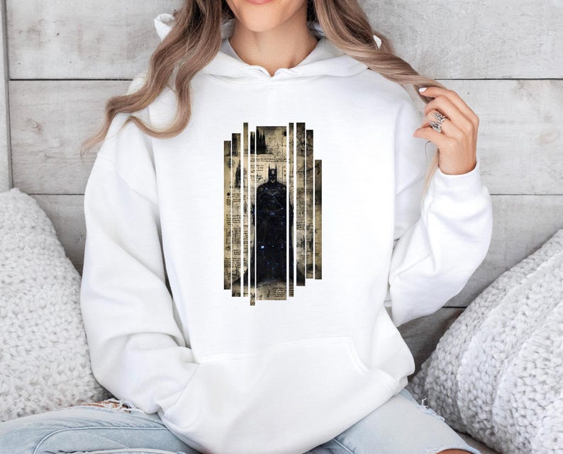 May include: White hoodie featuring a black Batman graphic. The graphic is a stylized image of Batman within a rectangular design. The design has a vintage newspaper background. The hoodie has a drawstring hood and a kangaroo pocket.