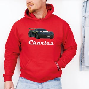 Puede incluir: Una sudadera con capucha roja con un dise&ntilde;o de coche de dibujos animados negro, con detalles verdes y amarillos. El nombre "Charles" est&aacute; impreso en blanco debajo del gr&aacute;fico del coche. La sudadera tiene un bolsillo delantero y una capucha con cord&oacute;n.