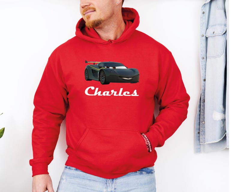 Puede incluir: Una sudadera con capucha roja con un dise&ntilde;o de coche de dibujos animados negro, con detalles verdes y amarillos. El nombre "Charles" est&aacute; impreso en blanco debajo del gr&aacute;fico del coche. La sudadera tiene un bolsillo delantero y una capucha con cord&oacute;n.