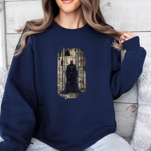 Puede incluir: Sudadera azul marino con un gr&aacute;fico de Batman. El gr&aacute;fico representa a Batman frente a un fondo de notas musicales y un paisaje urbano. La sudadera tiene mangas largas y cuello redondo.