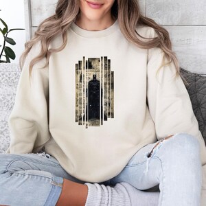 Puede incluir: Sudadera beige con un gr&aacute;fico de Batman. El dise&ntilde;o muestra una silueta negra de Batman dentro de un marco rectangular con un fondo de estilo peri&oacute;dico vintage. La sudadera es de color neutro.
