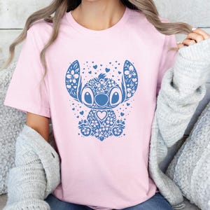 Puede incluir: Camiseta rosa claro con un dise&ntilde;o floral azul de un personaje de dibujos animados. El dise&ntilde;o incluye elementos florales, corazones y un detalle en forma de coraz&oacute;n. La camiseta es de cuello redondo.