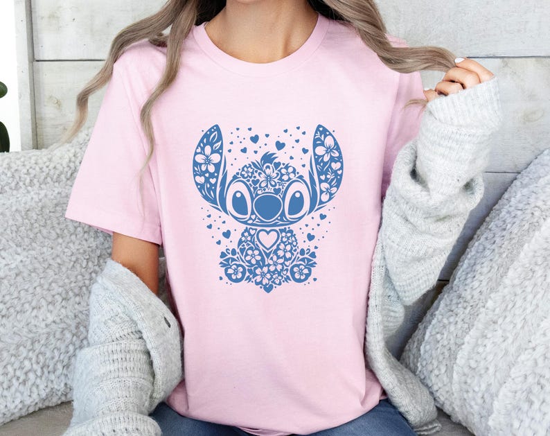 Puede incluir: Camiseta rosa claro con un dise&ntilde;o floral azul de un personaje de dibujos animados. El dise&ntilde;o incluye elementos florales, corazones y un detalle en forma de coraz&oacute;n. La camiseta es de cuello redondo.
