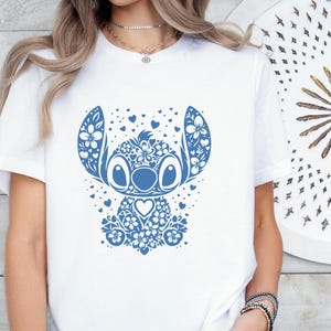 Puede incluir: Camiseta blanca con un dise&ntilde;o floral azul de un personaje de dibujos animados. El personaje tiene orejas grandes, ojos y una nariz en forma de coraz&oacute;n. El dise&ntilde;o incluye detalles florales y de coraz&oacute;n.