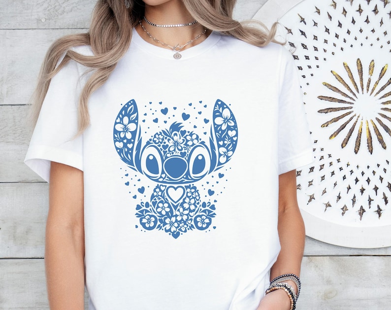 Puede incluir: Camiseta blanca con un dise&ntilde;o floral azul de un personaje de dibujos animados. El personaje tiene orejas grandes, ojos y una nariz en forma de coraz&oacute;n. El dise&ntilde;o incluye detalles florales y de coraz&oacute;n.