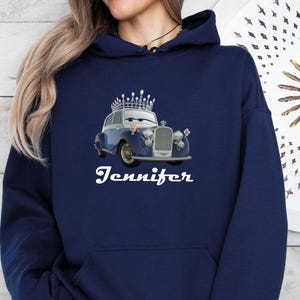 Puede incluir: Sudadera con capucha azul marino con un coche de dibujos animados con una corona. El coche es azul y plateado con un dise&ntilde;o vintage. El nombre "Jennifer" est&aacute; escrito en blanco debajo. La sudadera tiene un bolsillo delantero y una capucha con cord&oacute;n.