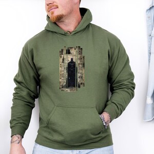 May include: Olive green hoodie with a black Batman graphic. The graphic is a stylized image of Batman in a rectangular frame. The hoodie has a front pocket and a drawstring hood. The sleeves are long.
