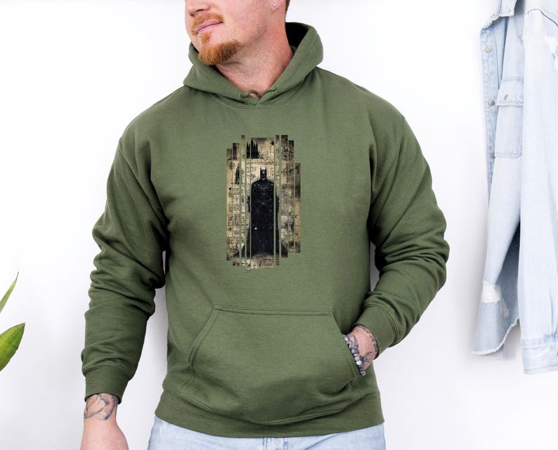 May include: Olive green hoodie with a black Batman graphic. The graphic is a stylized image of Batman in a rectangular frame. The hoodie has a front pocket and a drawstring hood. The sleeves are long.