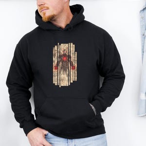 May include: Black hooded sweatshirt featuring a graphic of a figure with a red heart and red accents. The graphic is framed by vertical beige lines. The sweatshirt has a front pocket and a hood.