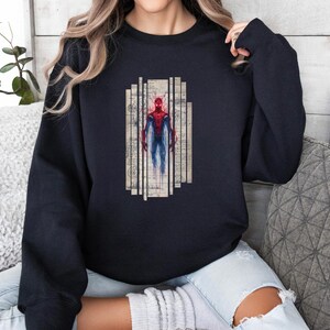 Op de afbeelding: Zwart sweatshirt met een Spiderman-afbeelding. De afbeelding toont Spiderman in een rood en blauw pak, tegen een vintage-achtige achtergrond met verticale lijnen. Het sweatshirt is effen zwart van kleur.