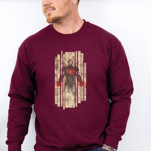 May include: Burgundy sweatshirt featuring a graphic of a superhero with a red glowing chest and hands, set against a vintage-style background. The design is framed by vertical lines. The sweatshirt is a solid burgundy color.