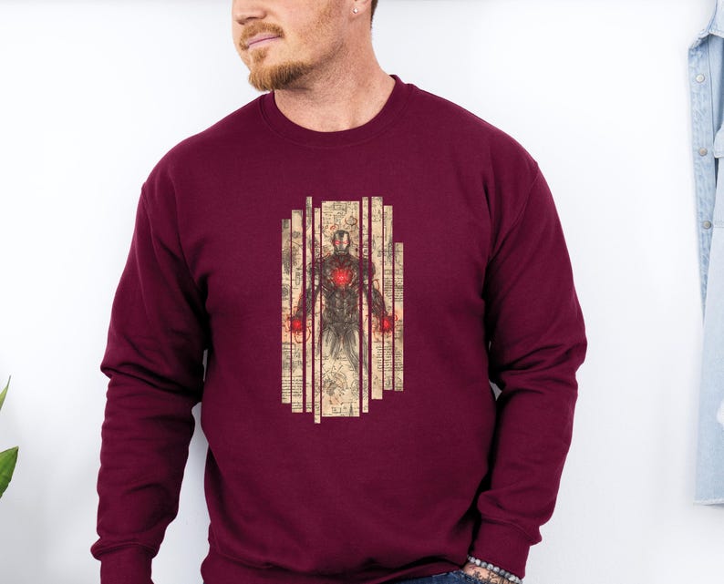 May include: Burgundy sweatshirt featuring a graphic of a superhero with a red glowing chest and hands, set against a vintage-style background. The design is framed by vertical lines. The sweatshirt is a solid burgundy color.