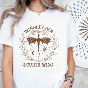 Pode incluir: Camiseta branca com um gr&aacute;fico marrom de um drag&atilde;o com as asas abertas, rodeado de estrelas e uma lua crescente. O texto "WINGLEADER RIDERS QUADRANT" est&aacute; acima do drag&atilde;o e "FOURTH WING" est&aacute; abaixo do drag&atilde;o.