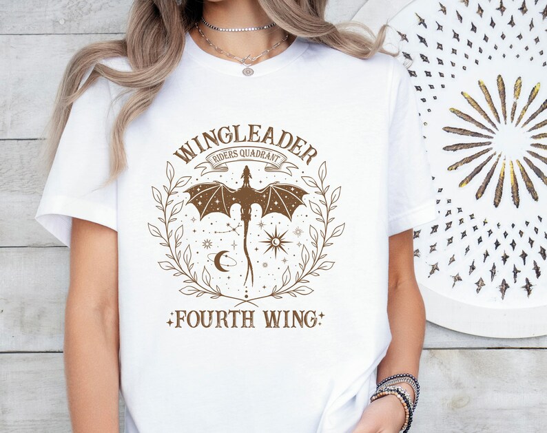 Pode incluir: Camiseta branca com um gr&aacute;fico marrom de um drag&atilde;o com as asas abertas, rodeado de estrelas e uma lua crescente. O texto "WINGLEADER RIDERS QUADRANT" est&aacute; acima do drag&atilde;o e "FOURTH WING" est&aacute; abaixo do drag&atilde;o.