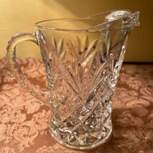 May include: A clear glass pitcher with a cut glass design. The pitcher has a wide mouth and a handle.
