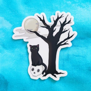May include: A black cat sits on a skull under a bare tree with a full moon in the background.