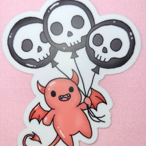 May include: A red cartoon devil with horns and wings holding three black and white skull balloons.