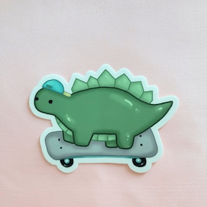 May include: A green dinosaur wearing a blue hat is riding a skateboard. The dinosaur has a spiky back and is facing to the right. The skateboard is grey with black wheels.