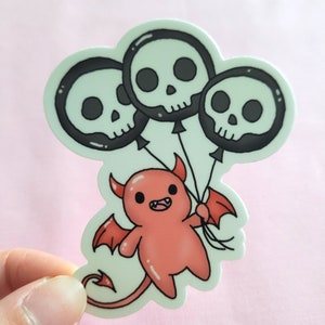 May include: A red cartoon devil with horns and wings is holding three black and white skull balloons. The balloons are tied together with a string.