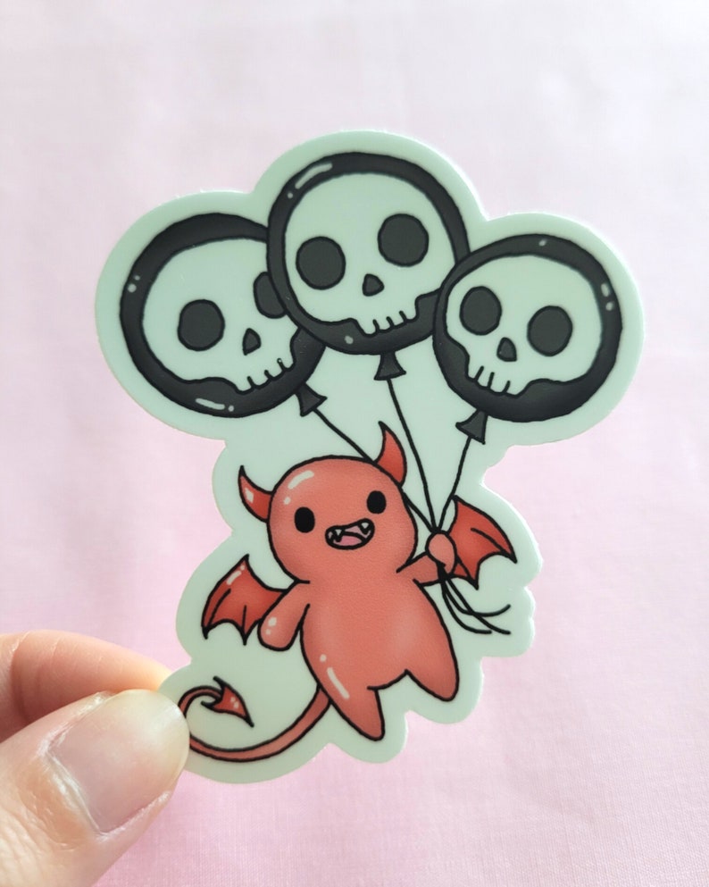 May include: A red cartoon devil with horns and wings is holding three black and white skull balloons. The balloons are tied together with a string.