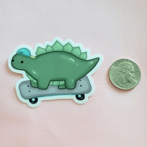 May include: A green dinosaur wearing a blue hat is skateboarding. The dinosaur has a spiky back and is on a grey skateboard with black wheels.