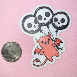May include: A red cartoon devil with wings and a tail holds three black and white skull balloons. The balloons have black eyes and a white outline.