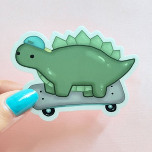 May include: A green dinosaur wearing a blue hat is skateboarding. The dinosaur has a spiky back and is on a grey skateboard with black wheels.
