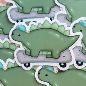 May include: A set of green dinosaur stickers with blue hats and grey skateboards. The dinosaurs are cartoon-style and have a friendly expression.