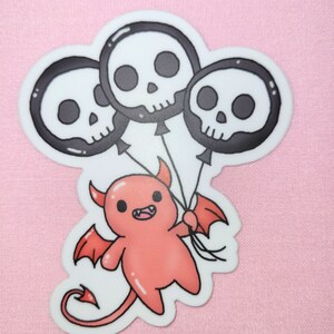 May include: A red cartoon devil with horns and wings holding three black and white skull balloons.