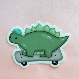 May include: A green dinosaur wearing a blue baseball cap is riding a skateboard with black wheels. The dinosaur has a spiky back and a long tail.