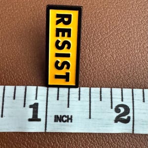 K&ouml;nnte beinhalten: Eine gelbe und schwarze Emaille-Anstecknadel mit dem Wort "RESIST" in Gro&szlig;buchstaben.
