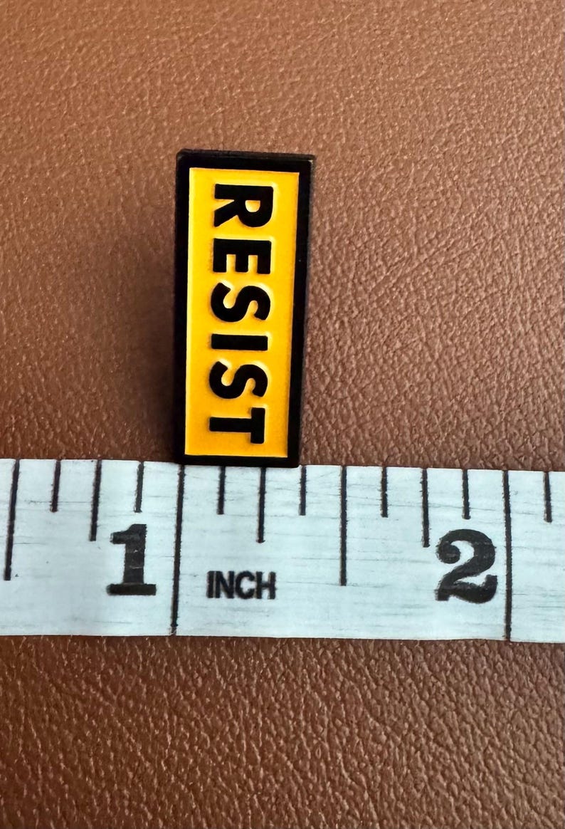 K&ouml;nnte beinhalten: Eine gelbe und schwarze Emaille-Anstecknadel mit dem Wort "RESIST" in Gro&szlig;buchstaben.
