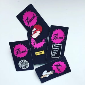 K&ouml;nnte beinhalten: F&uuml;nf schwarze und rosa Karten mit dem Wort "Flared" auf jeder. Die Karten zeigen verschiedene grafische Designs, darunter eine Person mit roten Haaren, eine Bergkette, ein Textfeld mit dem Wort "RESIST" und eine Namensliste.