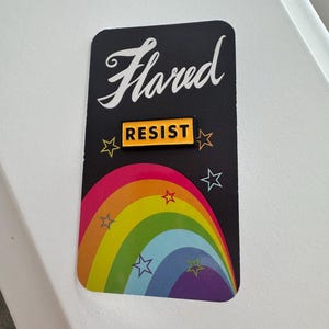 K&ouml;nnte beinhalten: Eine schwarze Karte mit dem Wort "Flared" in wei&szlig;er Kursivschrift. Ein gelber Knopf mit dem Wort "Resist" in schwarzen Buchstaben befindet sich auf der Karte. Ein Regenbogenbogen mit wei&szlig;en Sternen befindet sich hinter dem Knopf.