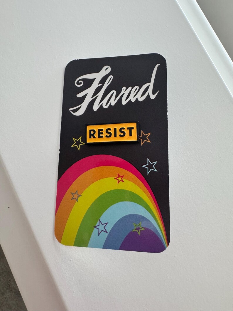 K&ouml;nnte beinhalten: Eine schwarze Karte mit dem Wort "Flared" in wei&szlig;er Kursivschrift. Ein gelber Knopf mit dem Wort "Resist" in schwarzen Buchstaben befindet sich auf der Karte. Ein Regenbogenbogen mit wei&szlig;en Sternen befindet sich hinter dem Knopf.