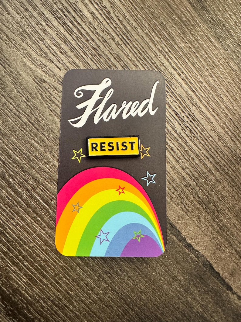 K&ouml;nnte beinhalten: Eine gelbe Emaille-Anstecknadel mit dem Wort "RESIST" in schwarzen Buchstaben auf schwarzem Hintergrund. Die Anstecknadel befindet sich auf einer Karte mit einem Regenbogen und Sternen.