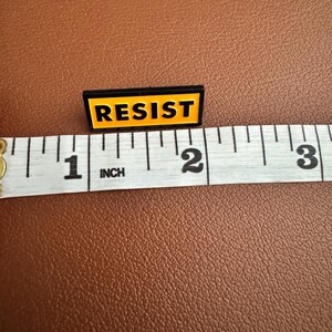 K&ouml;nnte beinhalten: Eine schwarze und gelbe Emaille-Anstecknadel mit dem Wort "RESIST" in Gro&szlig;buchstaben. Die Anstecknadel ist etwa 2,5 cm lang und liegt auf einer braunen Oberfl&auml;che neben einem Ma&szlig;band.