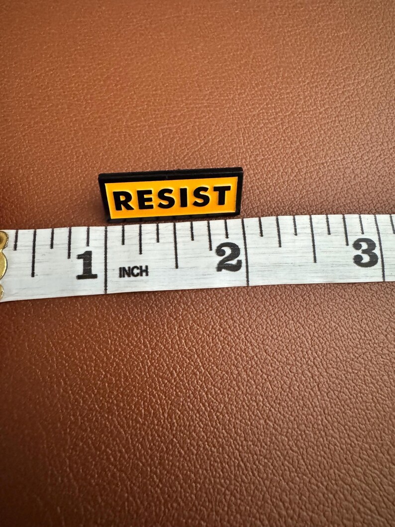 K&ouml;nnte beinhalten: Eine schwarze und gelbe Emaille-Anstecknadel mit dem Wort "RESIST" in Gro&szlig;buchstaben. Die Anstecknadel ist etwa 2,5 cm lang und liegt auf einer braunen Oberfl&auml;che neben einem Ma&szlig;band.