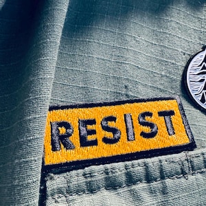 May include: Close-up of a green fabric with a yellow and black embroidered patch that reads "RESIST." The patch is rectangular and stitched onto the fabric. A small, circular emblem is visible in the upper right corner.