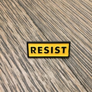 K&ouml;nnte beinhalten: Eine gelbe Emaille-Anstecknadel mit schwarzem Rand und dem Wort "RESIST" in schwarzen Buchstaben.