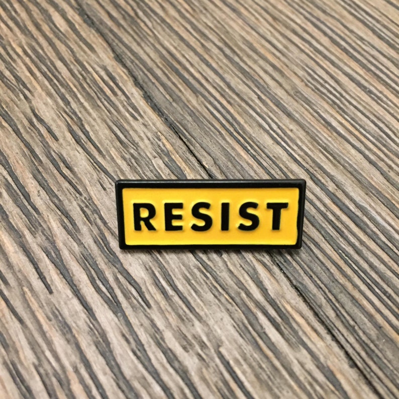K&ouml;nnte beinhalten: Eine gelbe Emaille-Anstecknadel mit schwarzem Rand und dem Wort "RESIST" in schwarzen Buchstaben.