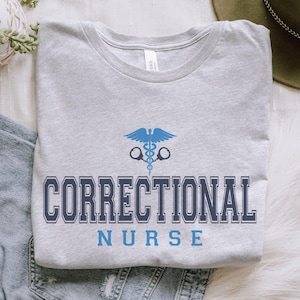 May include: A gray t-shirt with the text "Correctional Nurse" in blue lettering. A blue medical symbol with handcuffs is above the text.