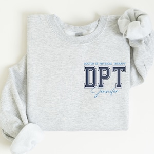 Puede incluir: Sudadera gris con el texto "Doctor of Physical Therapy DPT Jennifer" en letras azul marino y azul claro.